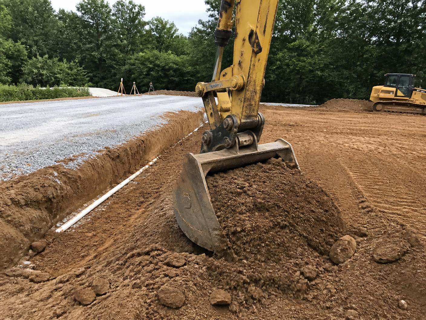Excavator bucket moving earth beside a fresh drainage trench and gravel pad
